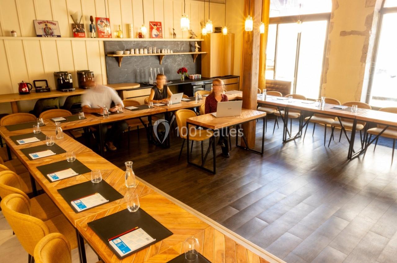 Salle de réunion lumineuse avec tables en U, chaises, matériel de travail et quelques personnes en activité.