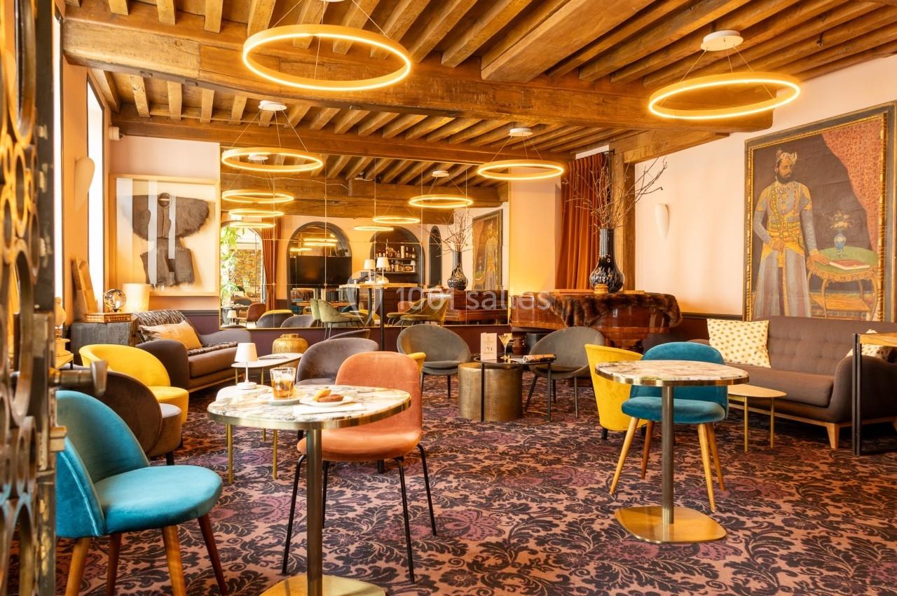 Salon chaleureux avec mobilier coloré, tables basses, éclairage suspendu circulaire et poutres apparentes au plafond.