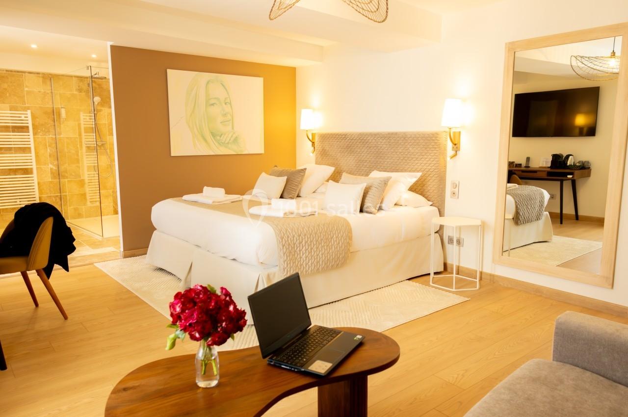 Chambre lumineuse avec lit double, salle de bain attenante, bureau avec ordinateur portable et vase de fleurs rouges.