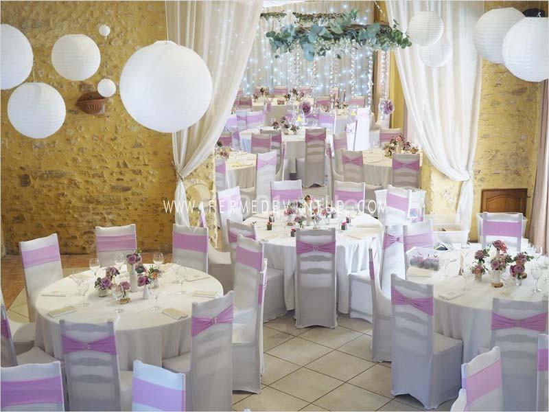 Salle de réception décorée avec des tables rondes, nappes blanches, chaises ornées de rubans roses et lanternes suspendues.