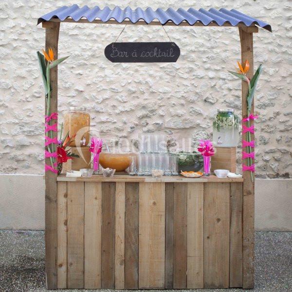Stand en bois avec des boissons, verres et décorations tropicales, sous une pancarte ’Bar à cocktail’.