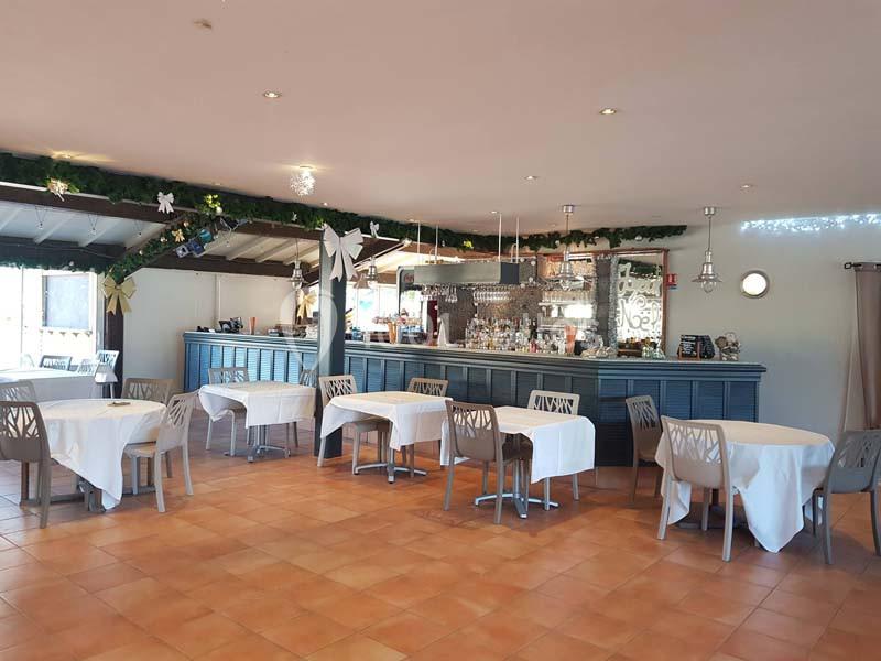 Salle de restaurant lumineuse avec tables dressées, chaises modernes et un bar décoré de guirlandes.