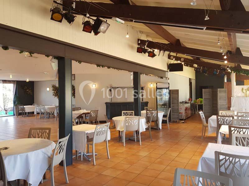 Salle de réception lumineuse avec tables rondes dressées, chaises modernes et poutres apparentes au plafond.