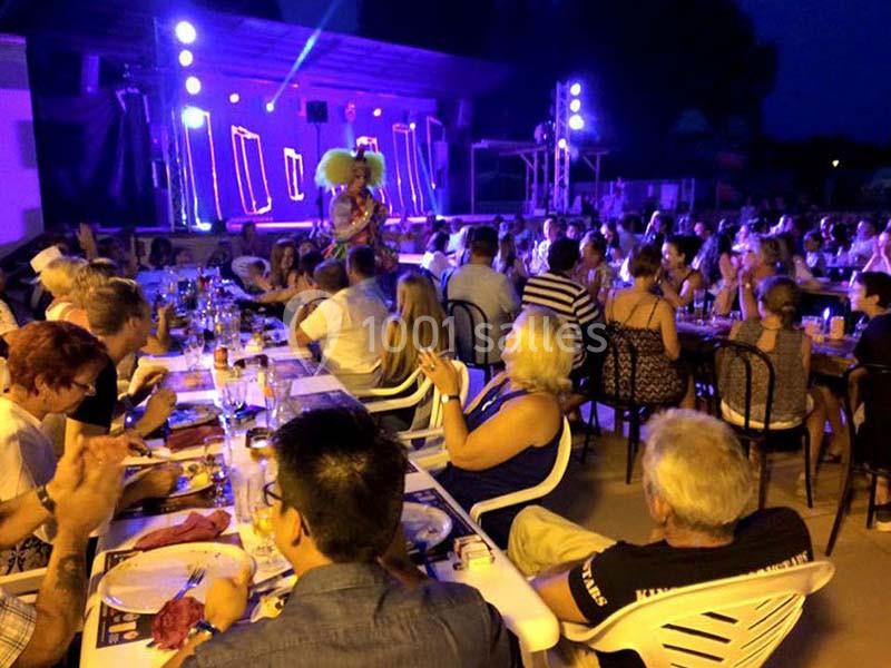 Groupe de personnes assises à des tables en extérieur, assistant à un spectacle sur une scène éclairée en soirée.