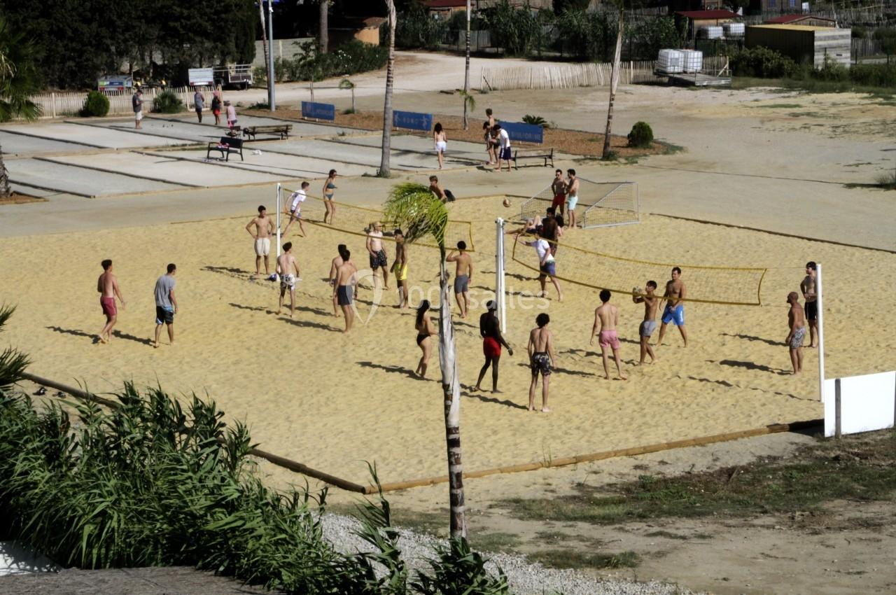Personnes jouant au volley-ball sur un terrain de sable en extérieur, entouré de végétation et d'aménagements urbains.