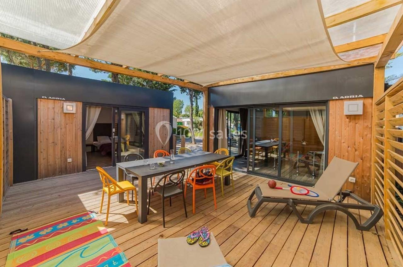 Terrasse en bois aménagée avec table, chaises colorées, transat et jouets, entourée de cabanes modernes en bois.