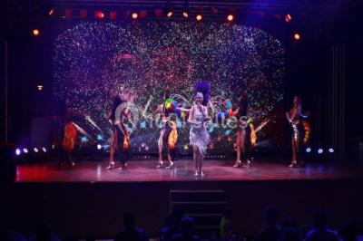 Groupe de personnes assises à des tables en extérieur, assistant à un spectacle sur une scène éclairée en soirée.