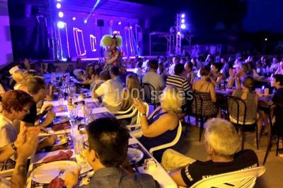 Groupe de personnes assises à des tables en extérieur, assistant à un spectacle sur une scène éclairée en soirée.