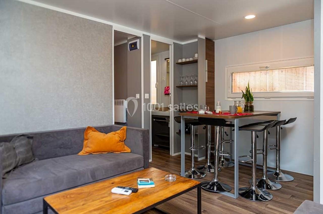 Salon d'un mobil-home avec canapé, table basse en bois, coin repas avec tabourets hauts et lumière naturelle.