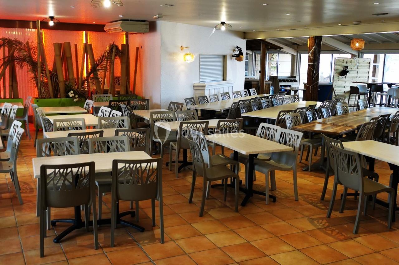 Salle de restaurant lumineuse avec des tables et chaises alignées sur un sol en carrelage, décorée de plantes et bois.