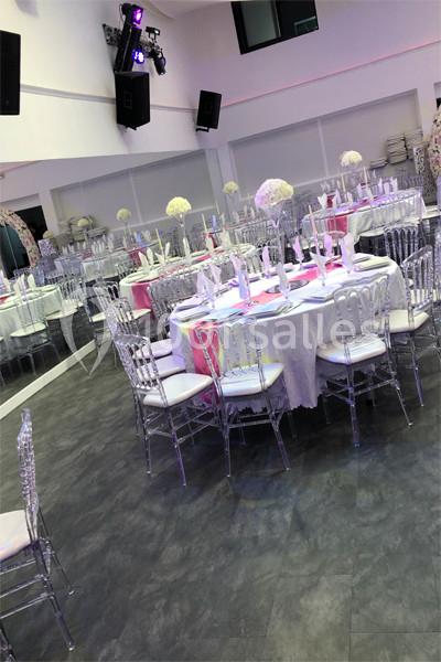 Salle de réception avec tables rondes dressées, nappes blanches, chaises transparentes et décorations florales.