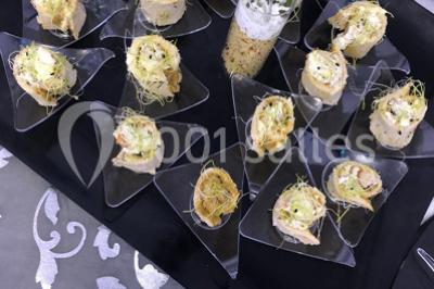 Cuillères transparentes disposées en cercle sur une assiette noire, remplies de semoule garnie de légumes et herbes.