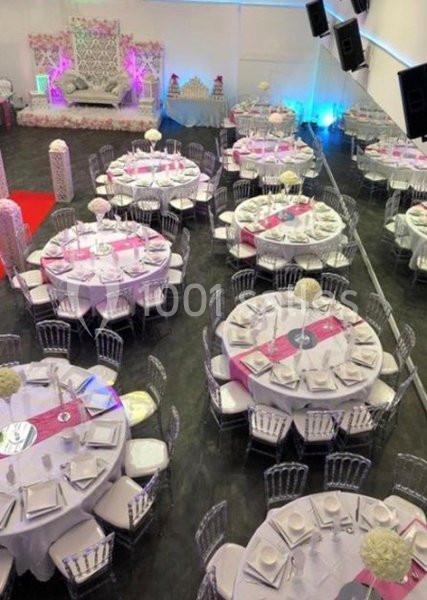 Salle de réception décorée avec des tables rondes dressées, nappes blanches, chemins de table roses et chaises transparentes.