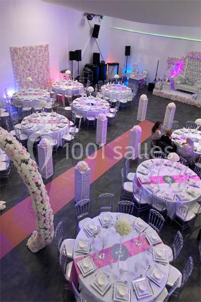 Salle de réception décorée avec des tables rondes, nappes blanches, chemins de table roses et colonnes florales.