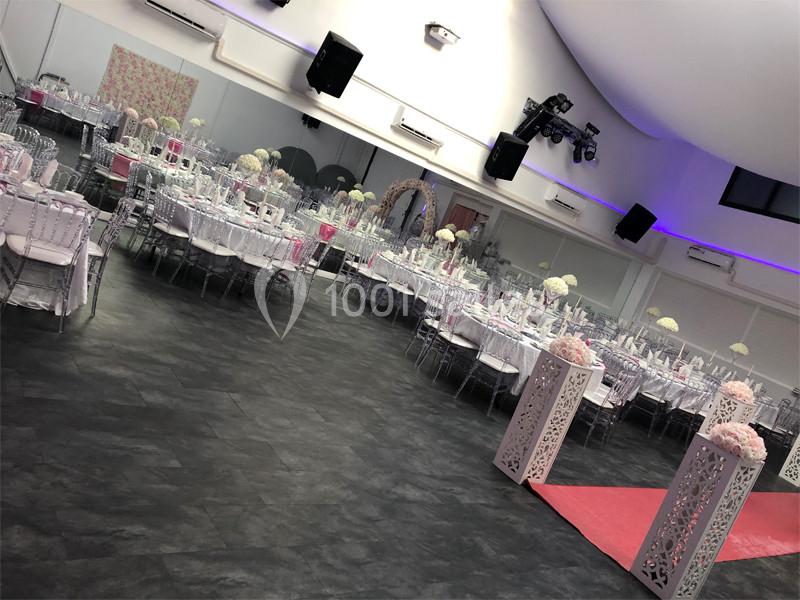 Salle de réception décorée avec des tables rondes, nappes blanches, chaises transparentes et allée rouge ornée de fleurs.