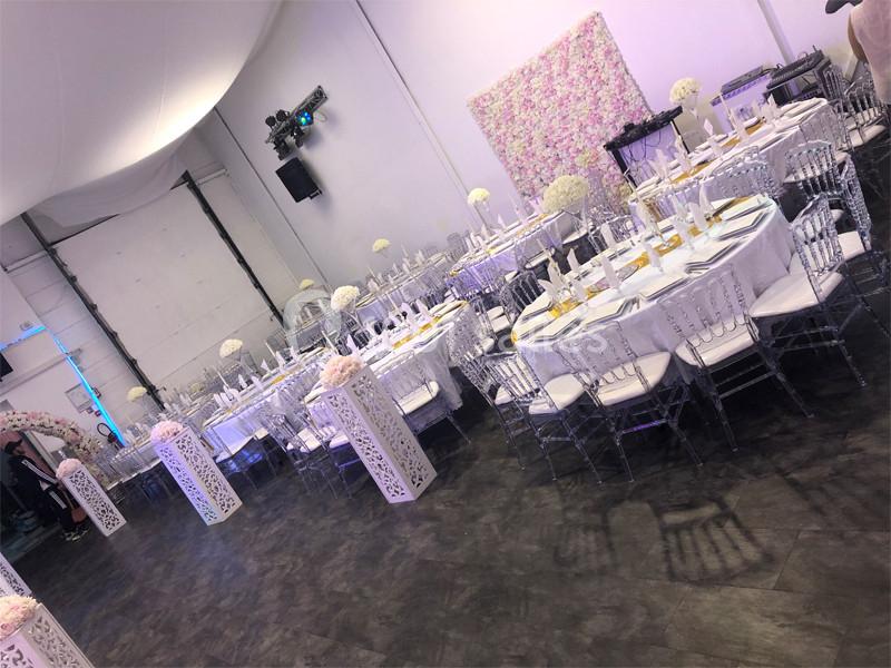 Salle de réception décorée avec des tables rondes, chaises transparentes et un mur floral en arrière-plan.
