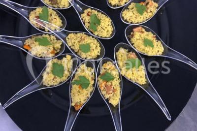 Cuillères transparentes disposées en cercle sur une assiette noire, remplies de semoule garnie de légumes et herbes.