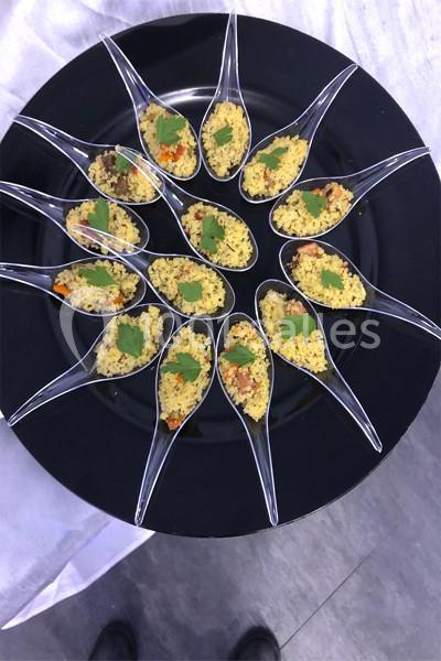 Cuillères transparentes disposées en cercle sur une assiette noire, remplies de semoule garnie de légumes et herbes.