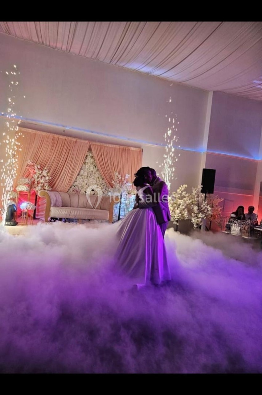 Un couple danse dans une salle décorée avec des fleurs, des rideaux roses et des effets lumineux sur une scène enfumée.
