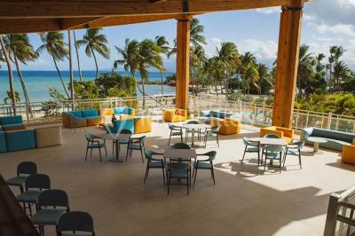 Plage bordée de palmiers avec eau turquoise, barrière rocheuse et montagnes à l'horizon sous un ciel dégagé.