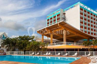 Plage bordée de palmiers avec eau turquoise, barrière rocheuse et montagnes à l'horizon sous un ciel dégagé.