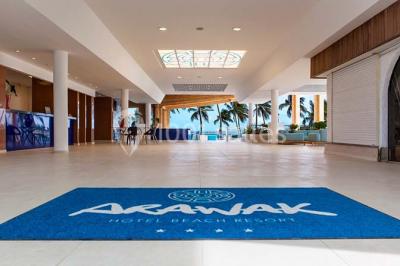 Plage bordée de palmiers avec eau turquoise, barrière rocheuse et montagnes à l'horizon sous un ciel dégagé.