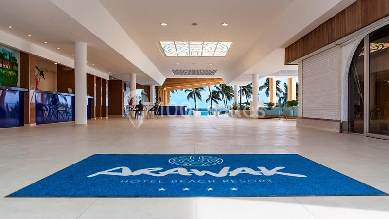 Entrée d'un hôtel avec un tapis bleu marqué ’Arawak’, vue sur un hall lumineux et des palmiers à l'extérieur.