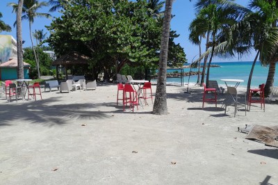 Terrasse couverte avec mobilier coloré, vue sur la mer turquoise et des palmiers sous un ciel dégagé.