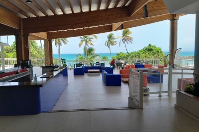 Terrasse couverte avec mobilier coloré, vue sur la mer turquoise et des palmiers sous un ciel dégagé.