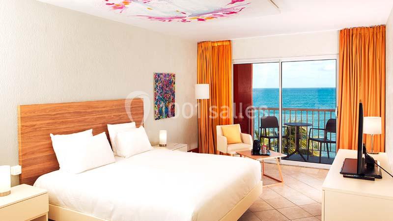 Chambre lumineuse avec lit double, balcon donnant sur la mer, rideaux orange et décoration colorée au mur.