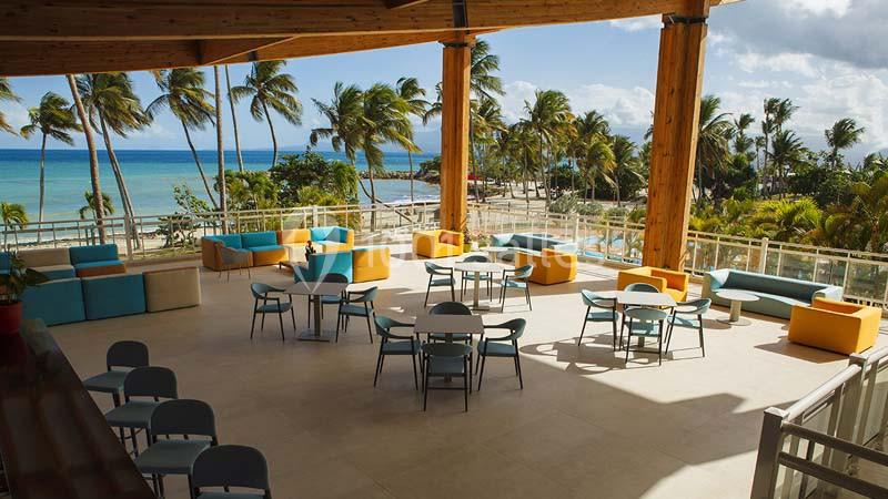 Terrasse aménagée avec tables, chaises et canapés colorés, offrant une vue sur la plage bordée de palmiers.