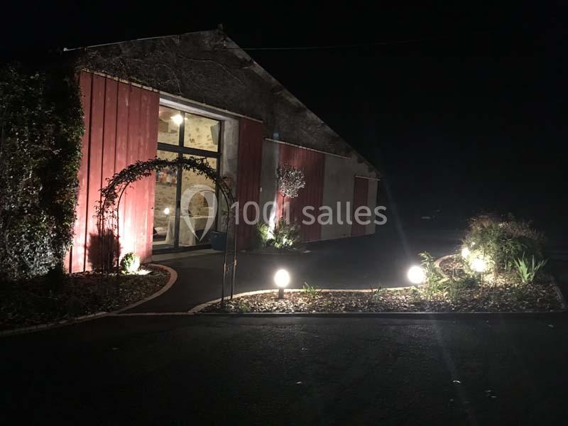 Entrée d'un bâtiment en pierre éclairée la nuit, avec des lumières et un jardin aménagé au premier plan.