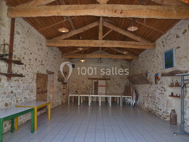 Salle en pierre avec poutres apparentes, tables et chaises empilées, éclairée par des lampes suspendues.