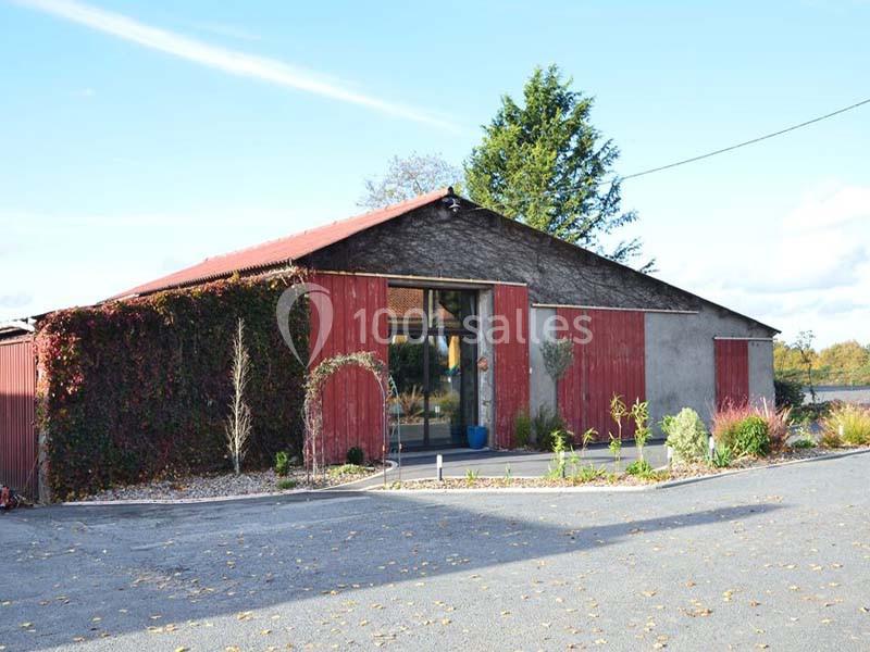 Bâtiment agricole rénové avec façade en bois rouge, entouré de végétation et d'un parking asphalté.