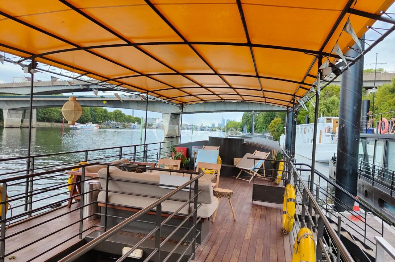 Pont d'un bateau aménagé avec des sièges sous une bâche jaune, amarré près d'un quai bordé d'arbres.