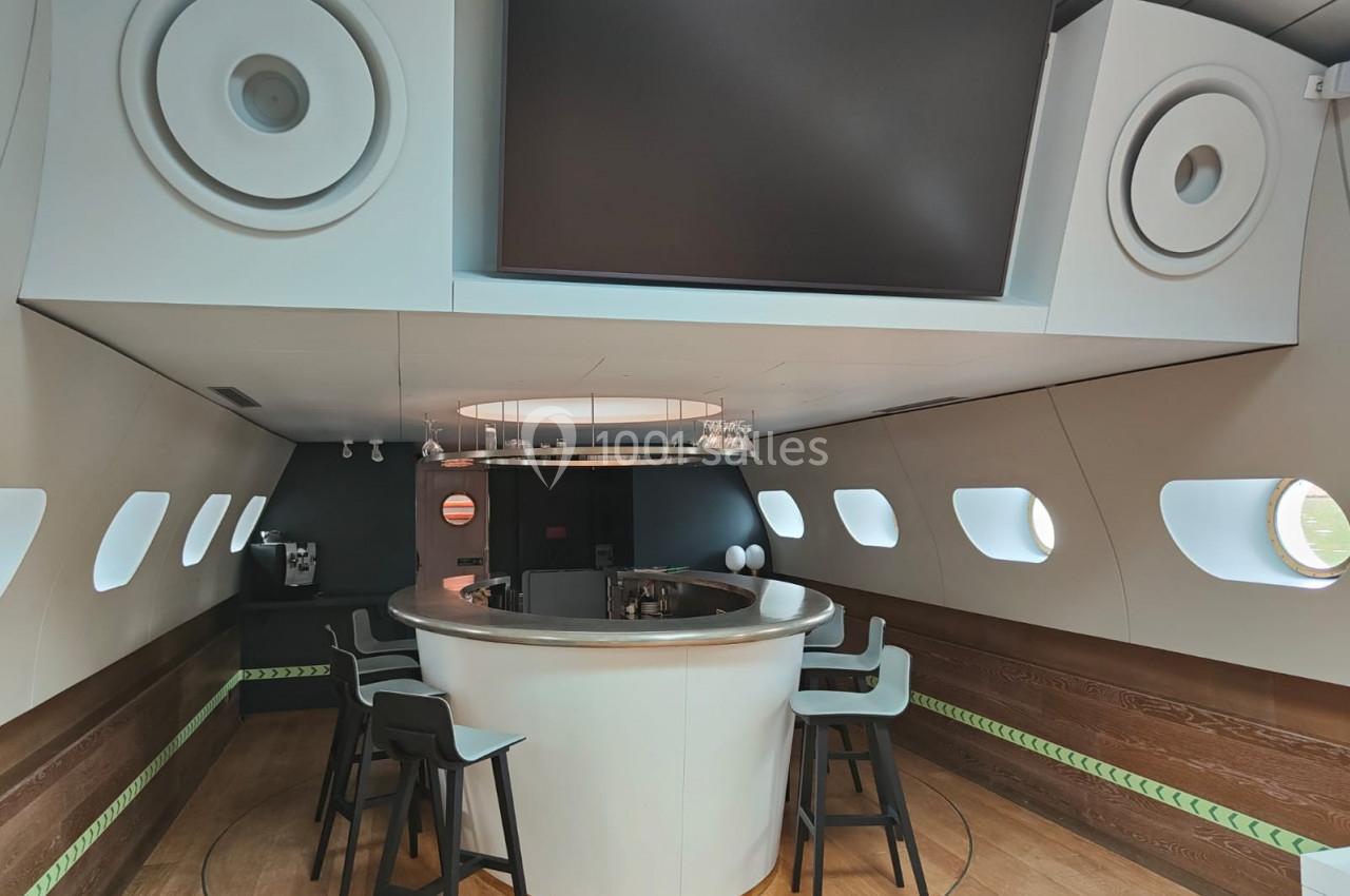 Bar circulaire moderne avec tabourets, situé dans un espace intérieur lumineux avec hublots et grands haut-parleurs intégrés.