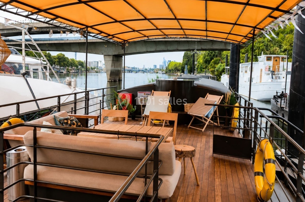Terrasse en bois d'un bateau avec canapés, chaises longues et toit jaune, amarré près d'autres bateaux sur une rivière.