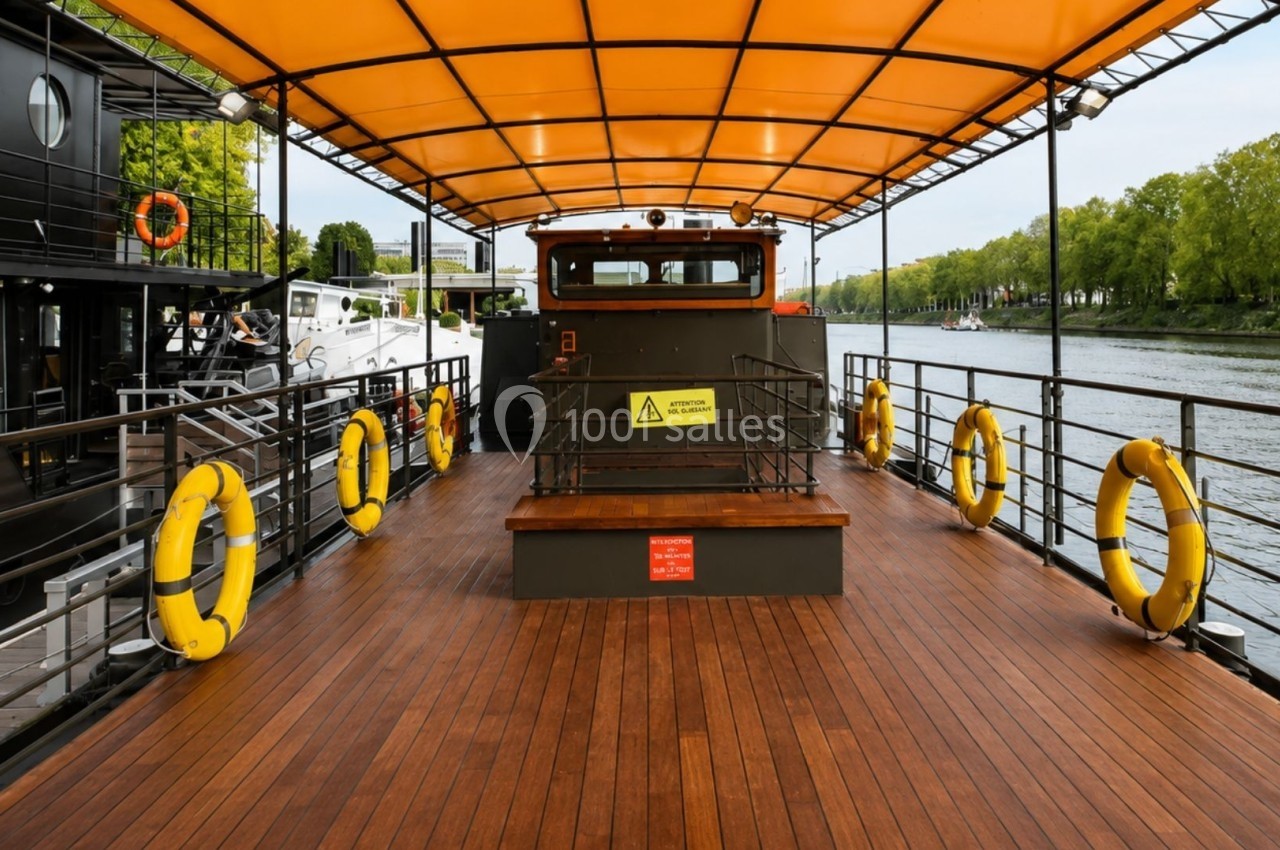 Pont en bois d'un bateau avec toit orange, bouées de sauvetage jaunes et vue sur une rivière bordée d'arbres.