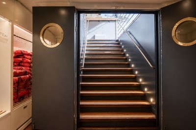 Escalier en bois éclairé menant à l'extérieur, encadré par des portes grises avec hublots circulaires.