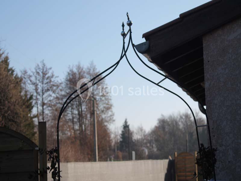 Arche métallique décorative dans un jardin, près d'une maison, avec des arbres et un ciel dégagé en arrière-plan.
