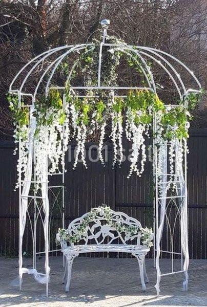 Structure en métal blanc ornée de plantes vertes et de fleurs blanches, avec un banc assorti en dessous.