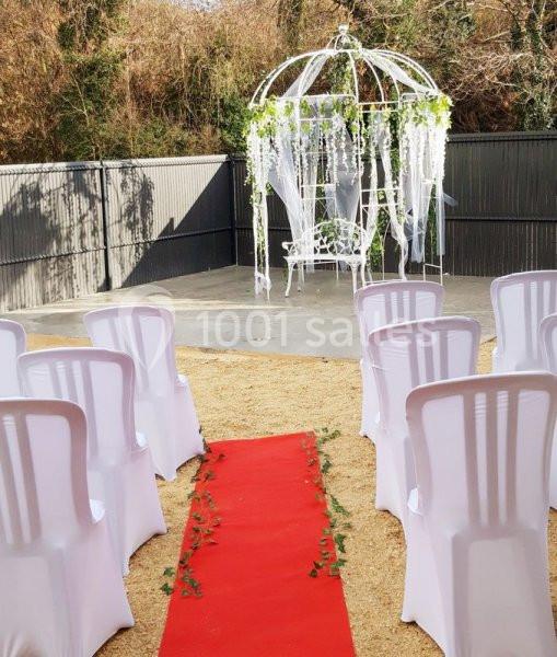 Chaises blanches alignées devant une arche décorée de voilages et de fleurs, avec un tapis rouge au centre.