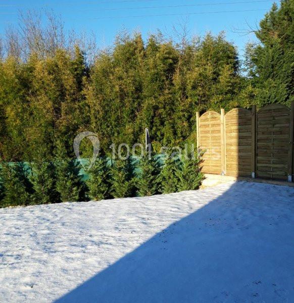 Pelouse enneigée bordée de haies de conifères et d'une clôture en bois sous un ciel dégagé.
