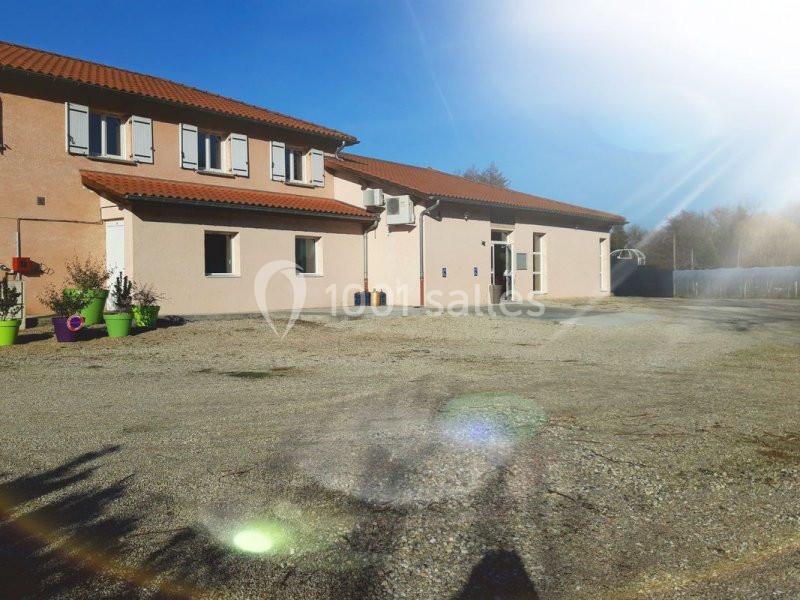 Bâtiment beige à deux étages avec toit en tuiles rouges, entouré d'une cour en gravier et de quelques plantes en pots.