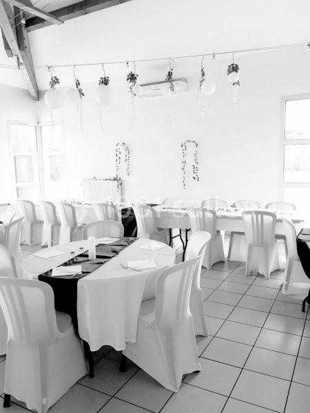 Salle décorée avec des tables et chaises blanches, nappes et guirlandes suspendues, éclairée par la lumière naturelle.