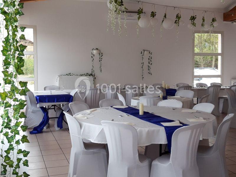 Salle décorée pour un événement, avec des tables rondes nappées de blanc et bleu, entourées de chaises blanches.