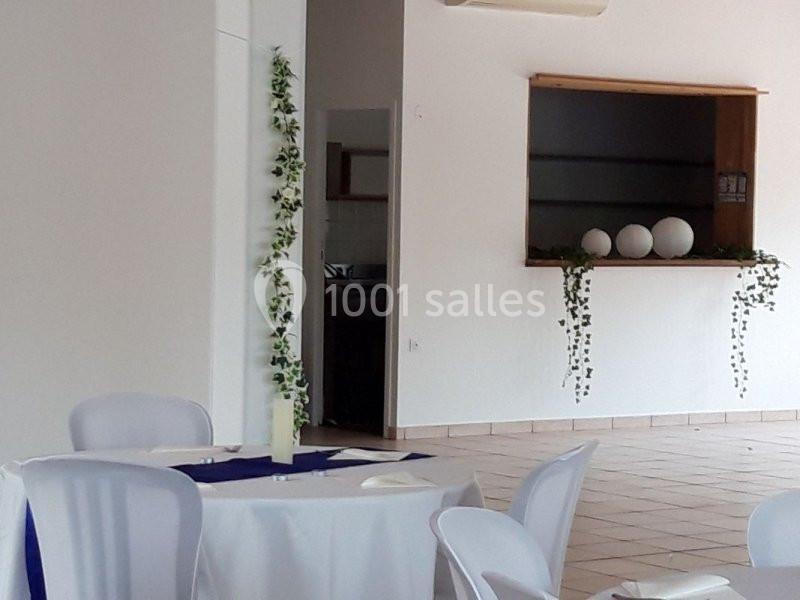 Salle lumineuse avec table ronde dressée, chaises blanches, décorations végétales et étagère murale en arrière-plan.