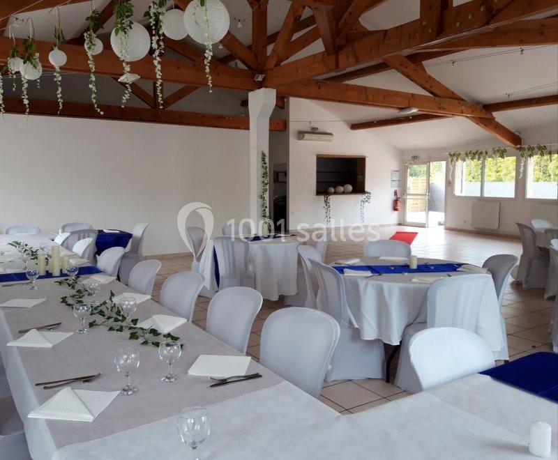 Salle de réception lumineuse avec tables dressées, nappes blanches, décorations végétales et poutres apparentes en bois.