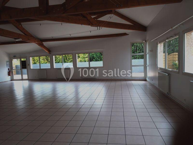 Salle vide avec carrelage clair, poutres apparentes au plafond, grandes fenêtres laissant entrer la lumière naturelle.