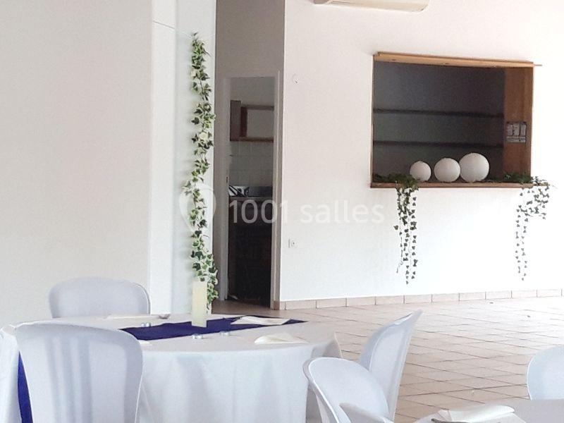 Salle lumineuse avec table ronde dressée, chaises blanches et décoration végétale sur un comptoir en arrière-plan.